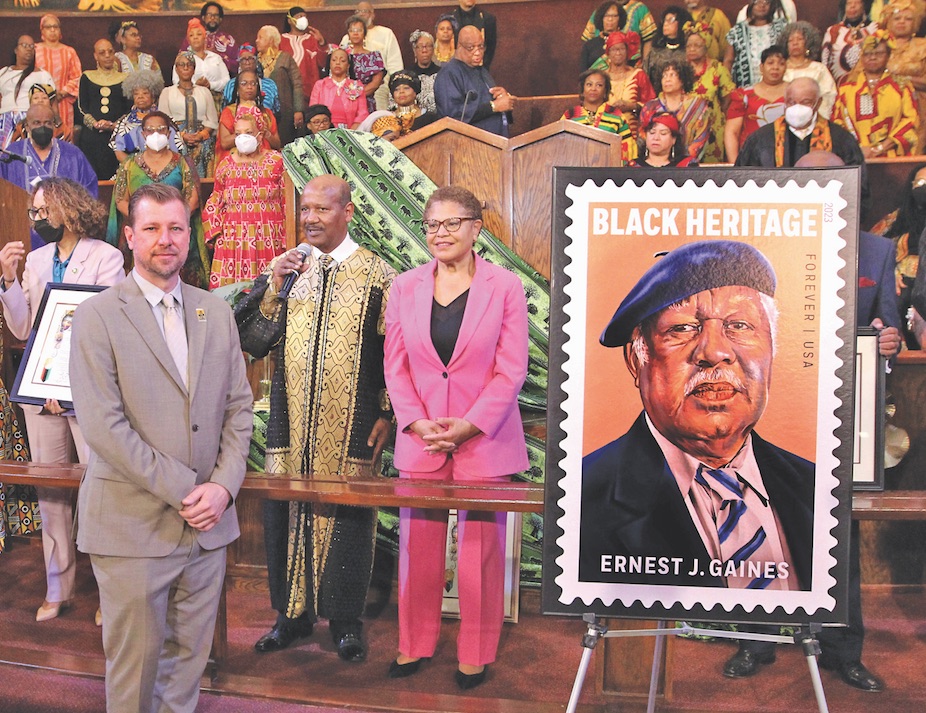 The image shows a group of people standing in front of a picture of a man. The text on the picture mentions "2023 Black Heritage Forever | USA Ernest J. Gaines." One of the individuals in the group is mayor Karen Bass, wearing a pink suit.